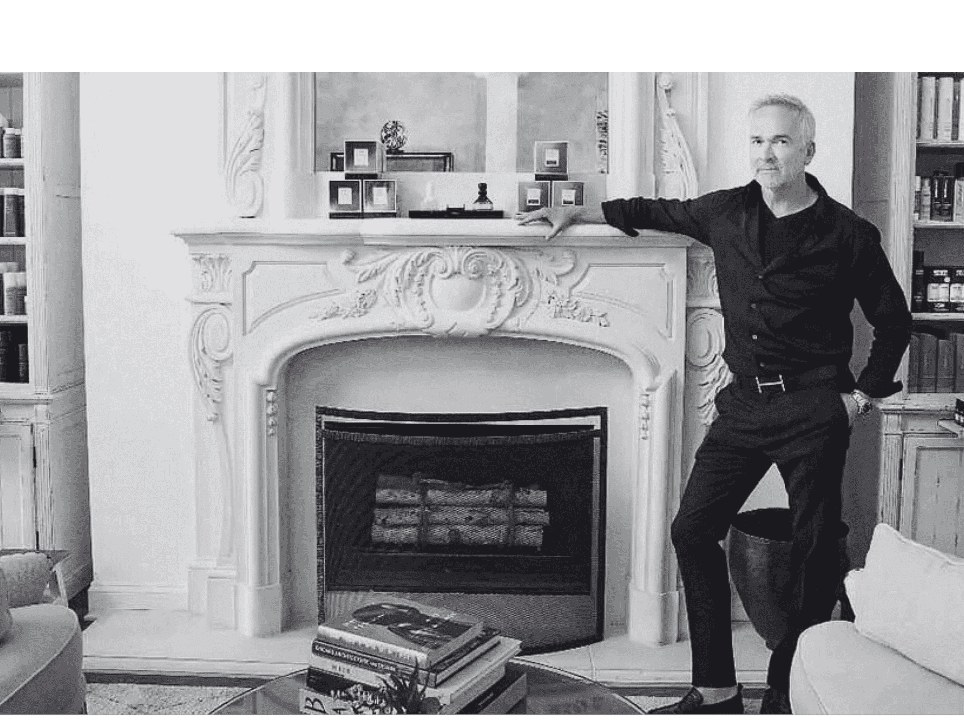 Man in black attire standing by an ornate fireplace, with shelves in the background.