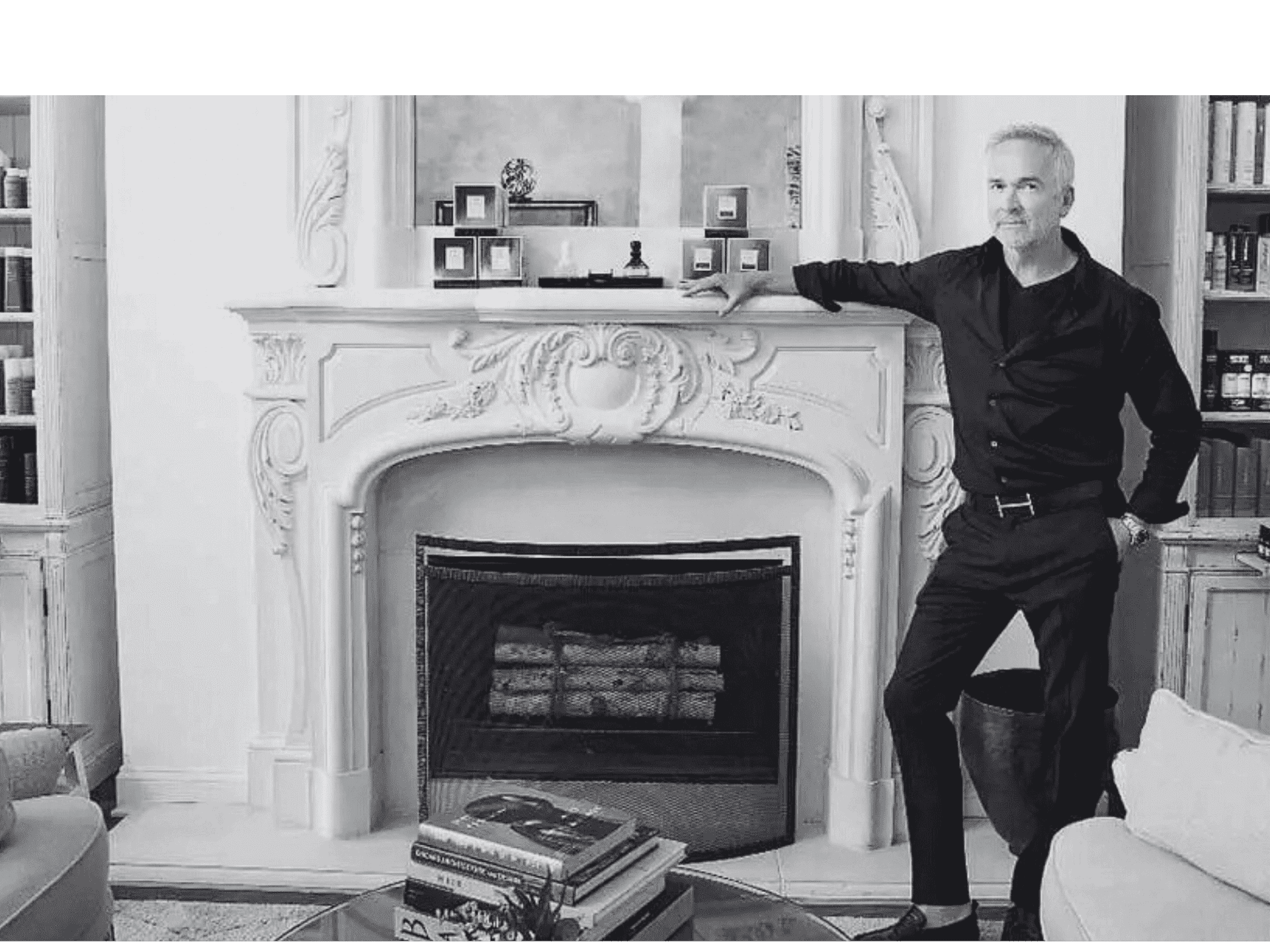 Man in black attire standing by an ornate fireplace, with shelves in the background.